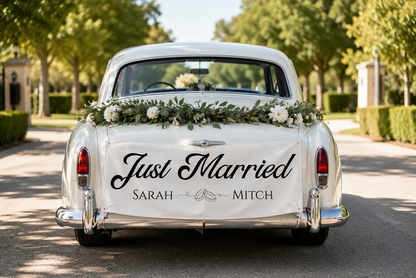 Rear view of a vintage white car on a tree-lined road, decorated with a green floral garland across the trunk and a large white banner reading 'Just Married' with the names 'Sarah' and 'Mitch'.