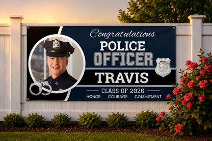 A personalized police officer graduation banner displayed on a white fence in a landscaped yard. The banner features a dark navy blue background with white and silver text reading 'Congratulations POLICE OFFICER TRAVIS CLASS OF 2026' and 'HONOR COURAGE COMMITMENT'.