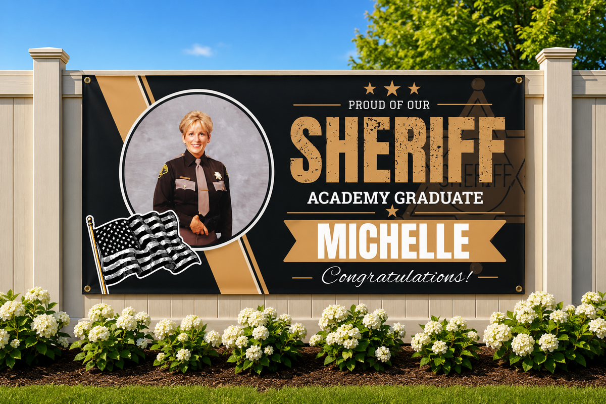 A large custom graduation banner mounted on a white fence, celebrating a sheriff academy graduate named Michelle. The banner features a black background with gold and beige graphic elements, a portrait of a female sheriff, an American flag graphic, and the text 'PROUD OF OUR SHERIFF ACADEMY GRADUATE MICHELLE Congratulations!'.