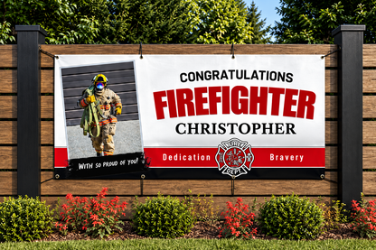 A personalized firefighter congratulations banner displayed on a wooden fence outdoors. The banner features a photo of a firefighter in full gear on the left, with the text 'CONGRATULATIONS FIREFIGHTER CHRISTOPHER' in black and red lettering on a white background.